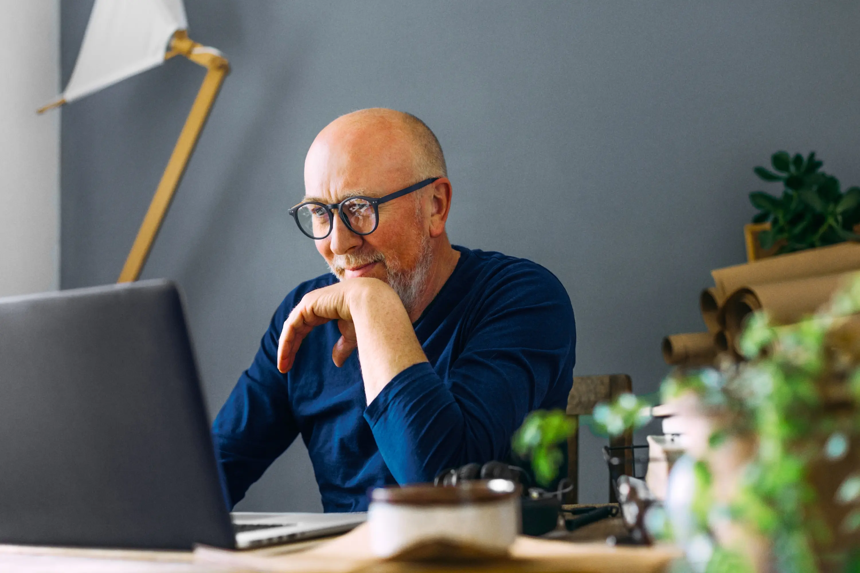 Raisin IE-Senior man with glasses using laptop A bald senior man with glasses and a grey beard leaning on his hand while looking thoughtfully at a laptop screen.