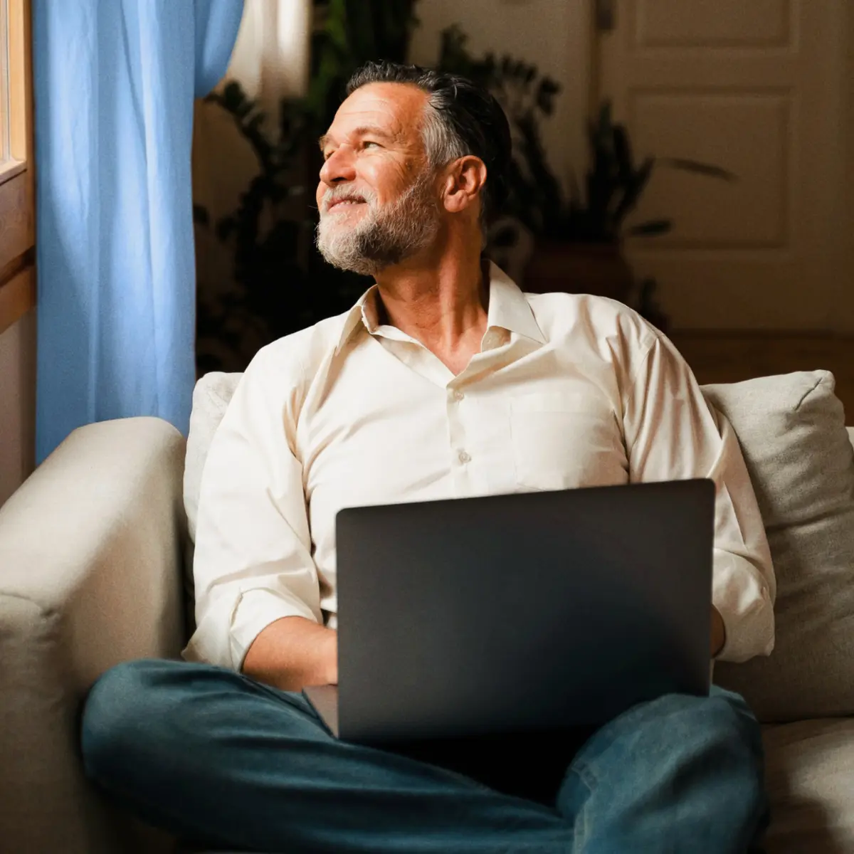 A smiling mature man sitting on a sofa with a laptop on his lap, looking out a window with light blue curtains.