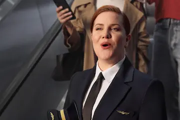 Eine Frau in Uniform steht in einem öffentlichen Innenraum und hält ein Smartphone. Sie blickt nach vorne und wirkt konzentriert. Im Hintergrund sind weitere Personen unscharf zu erkennen.