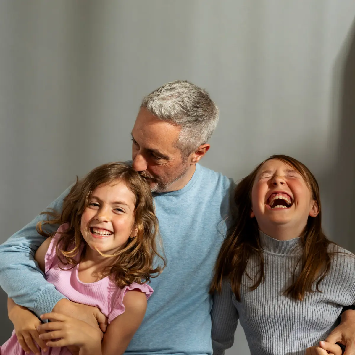 raisin_de_vater_kinder_lachen.png Vater umarmt zwei fröhlich lachende Kinder vor grauem Hintergrund.