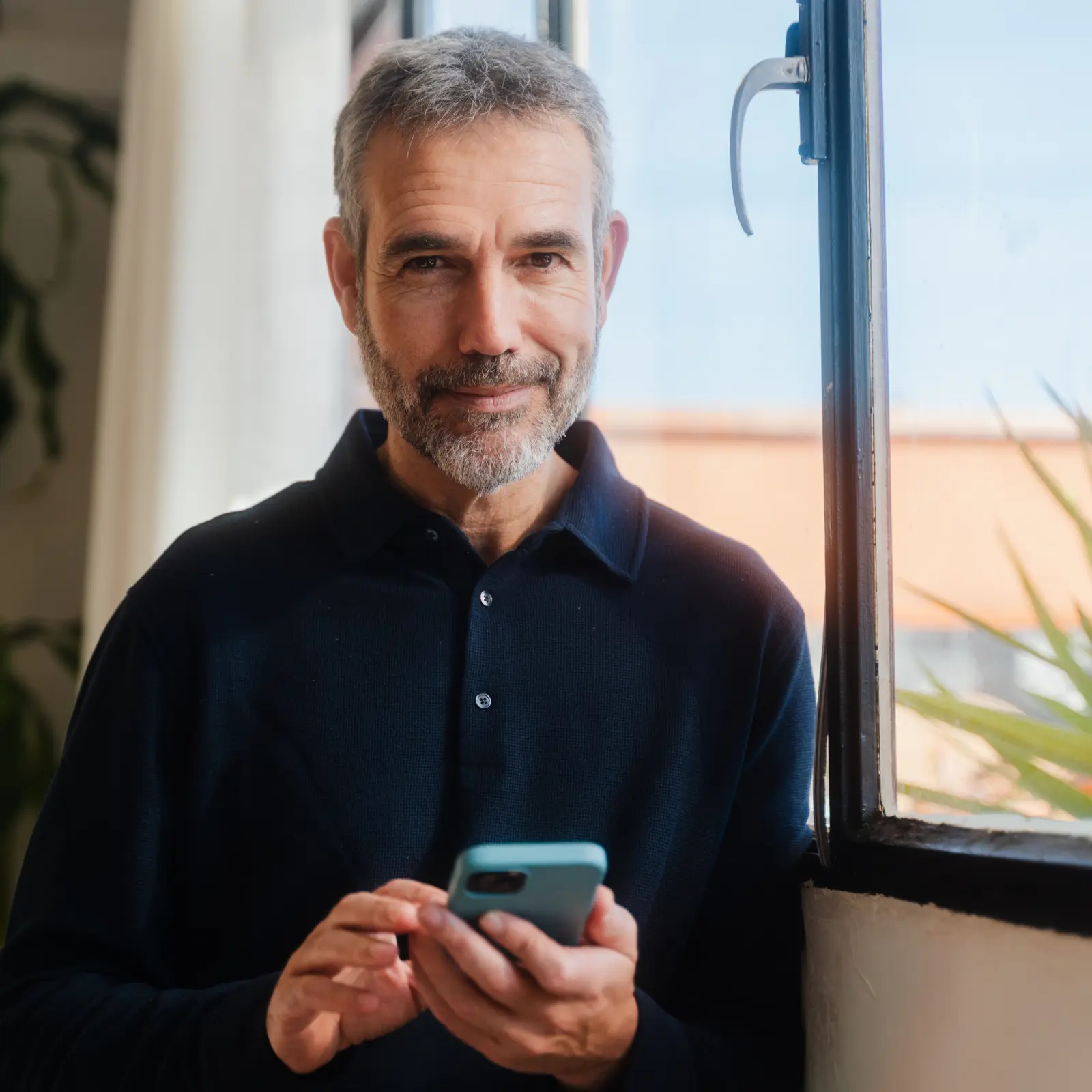 raisin_de_mann_smartphone_fenster.png Mann mit Brille lacht während eines Telefonats in einer hellen Küche und hält eine Tasse.