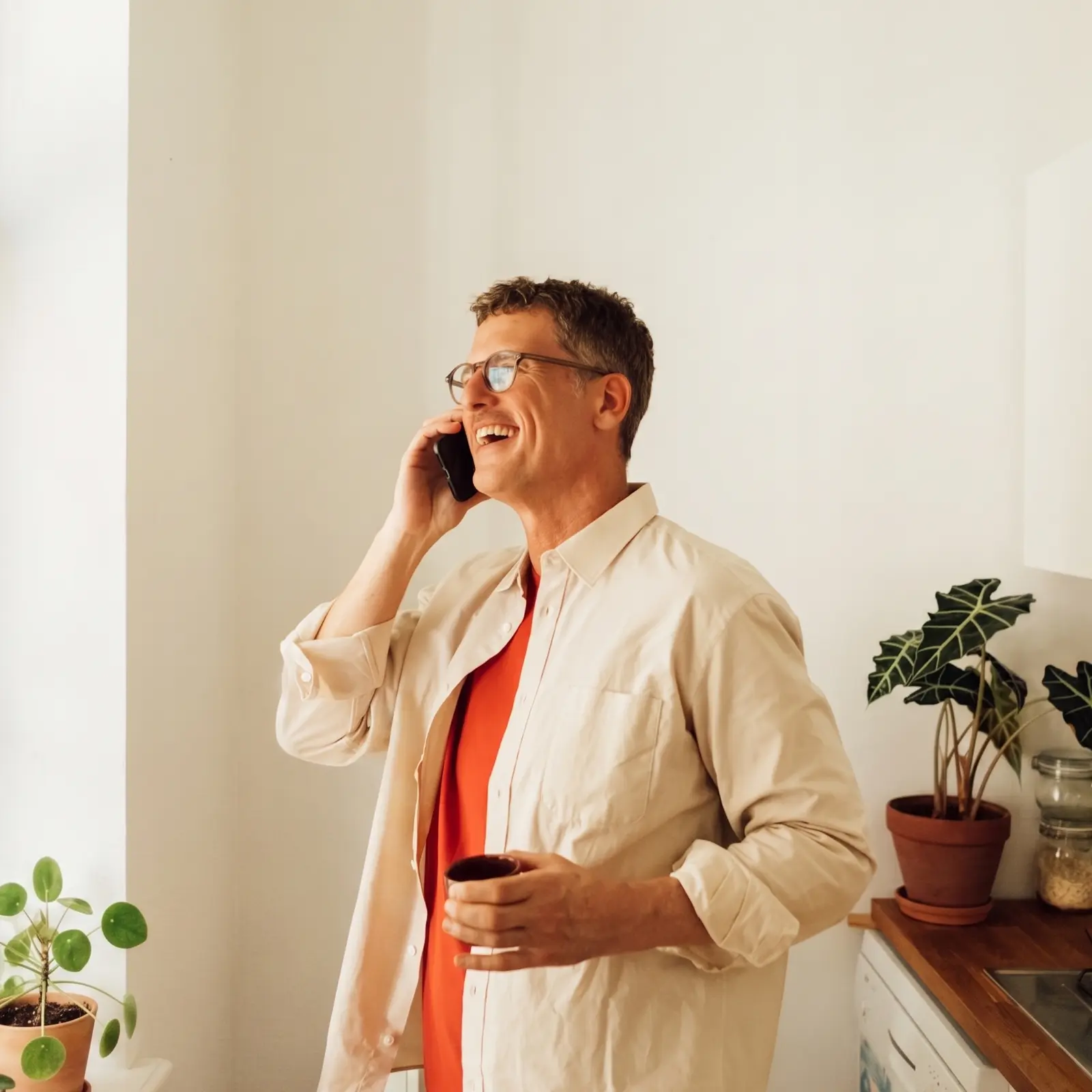 Mann mit Brille lacht während eines Telefonats in einer hellen Küche und hält eine Tasse.