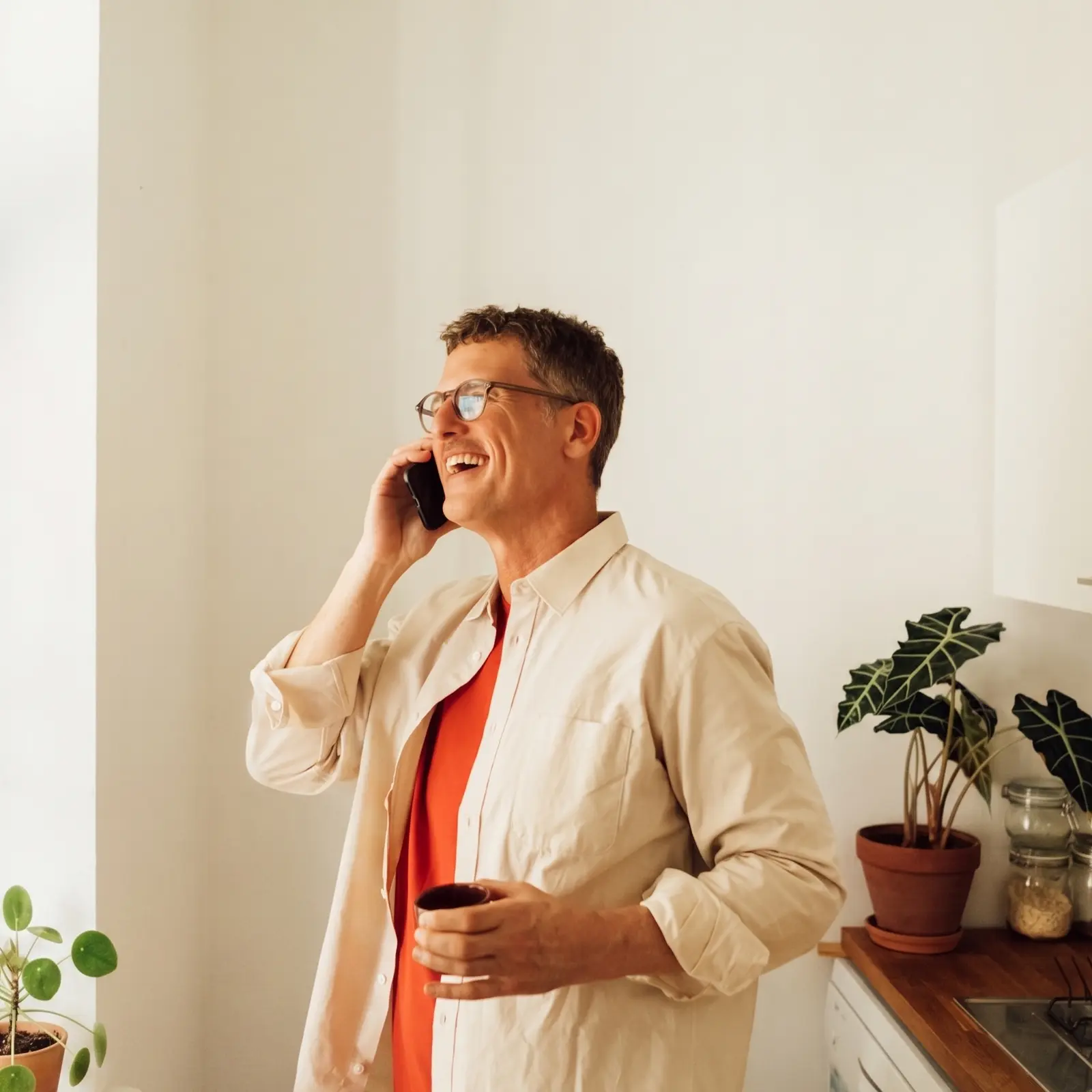 Ein Mann mit Brille lacht herzlich in der Küche, während er mit dem Smartphone telefoniert.