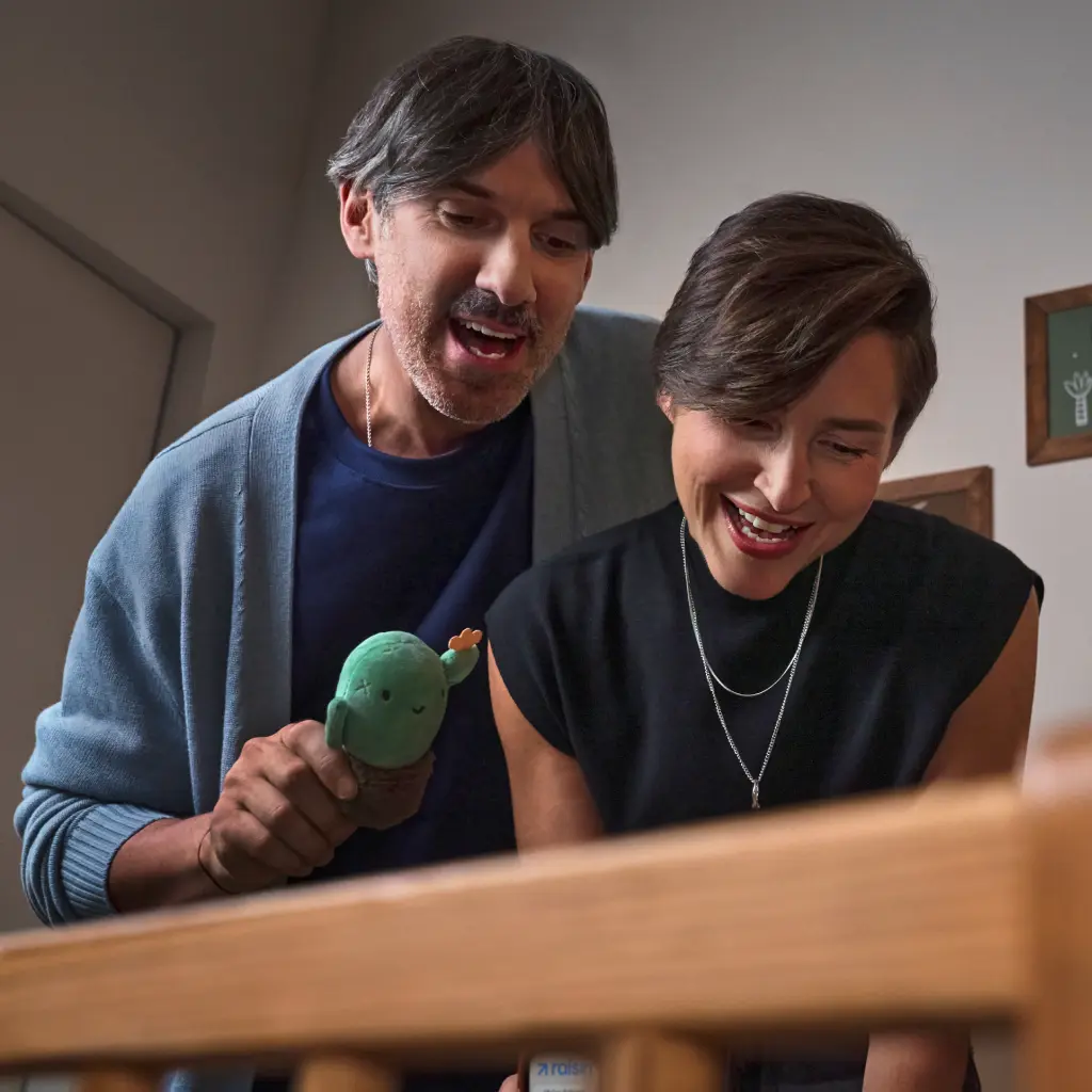 Raisin DE Kinderdepot Warteliste Zwei Erwachsene stehen nebeneinander und blicken lächelnd nach unten. Eine Person hält ein kleines grünes Stoffspielzeug in der Hand. Die Szene wirkt vertraut und verspielt, aufgenommen in einem wohnlichen Innenraum.