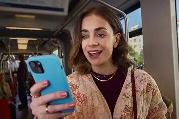 Eine Frau sitzt in einer Straßenbahn und schaut lächelnd auf ihr Smartphone. Sie hält das Gerät in der Hand, im Hintergrund sind weitere Fahrgäste und das Innere der Bahn zu sehen.