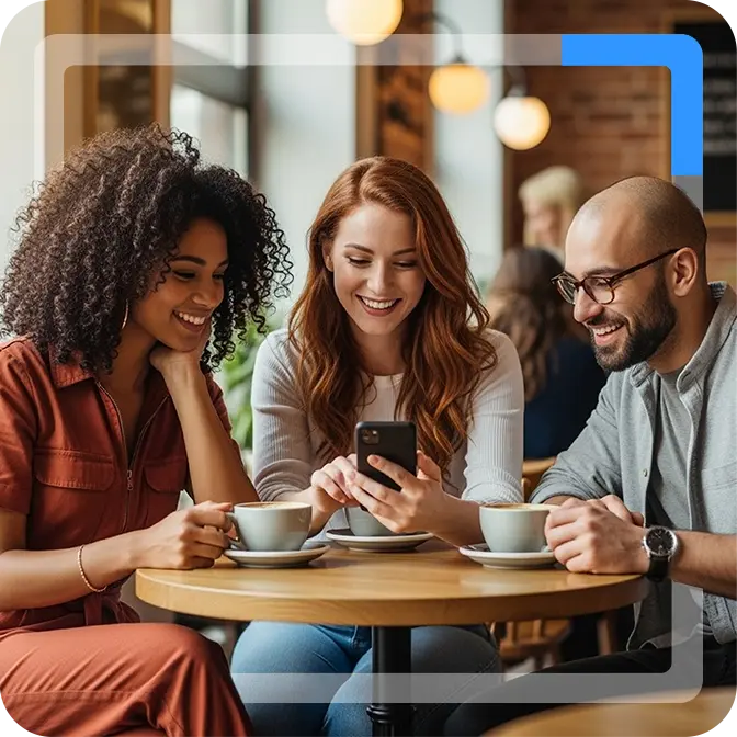 A group of three friends gathered around a coffee shop table. The person in the middle is holding a phone and showing her friends what is on the screen.