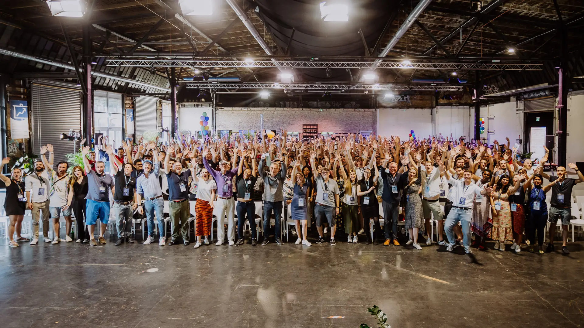 Wide group photo of several hundred Raisin employees in a large hall, standing in rows with their arms raised, smiling towards the camera.